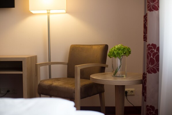 Una habitación de hotel con una cama, una silla de madera, una mesita con un florero de plantas verdes y una lámpara de pie.