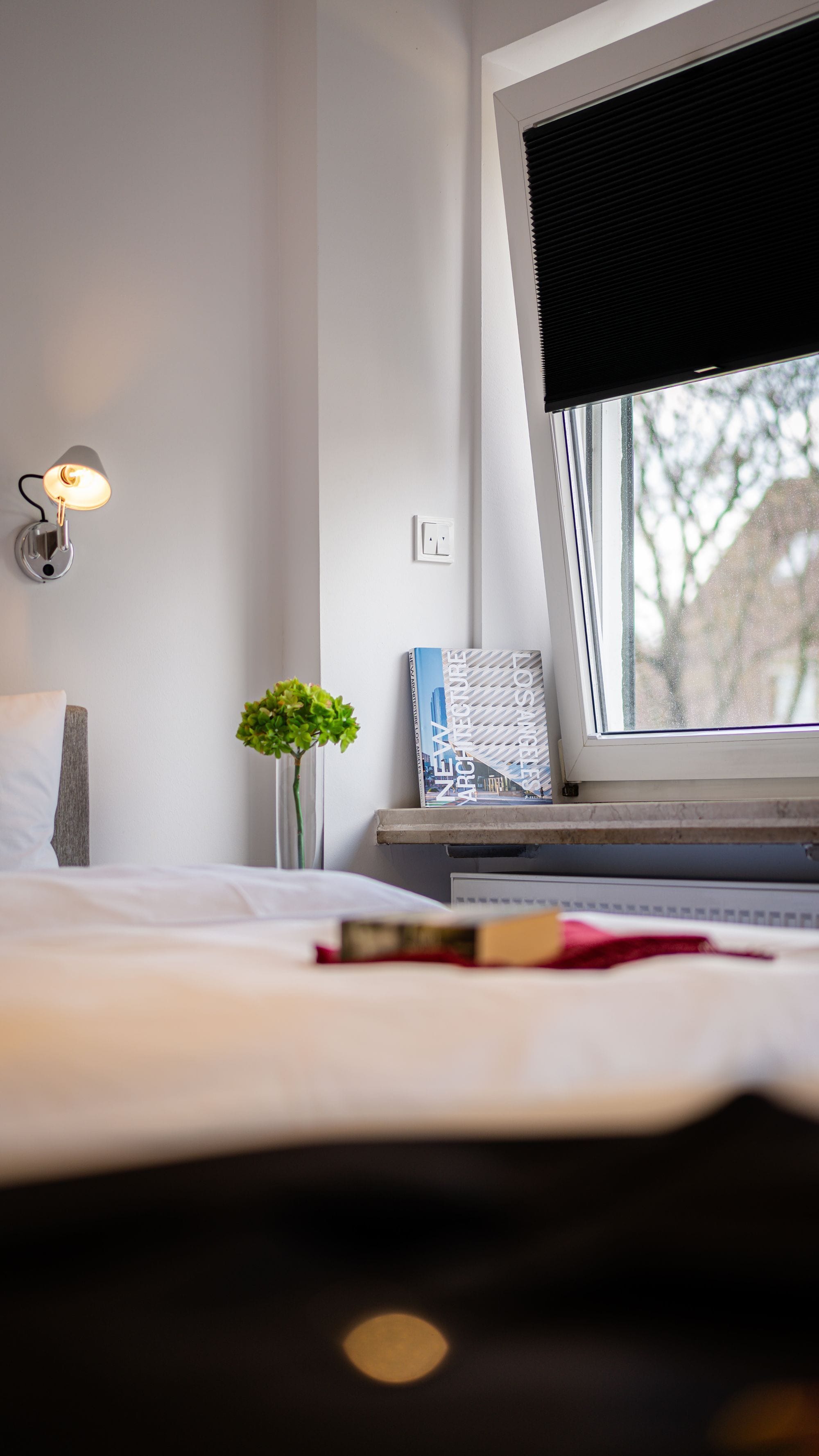 A bedroom with a bed, a bedside lamp, a small plant, a book, and a window with a view of trees.