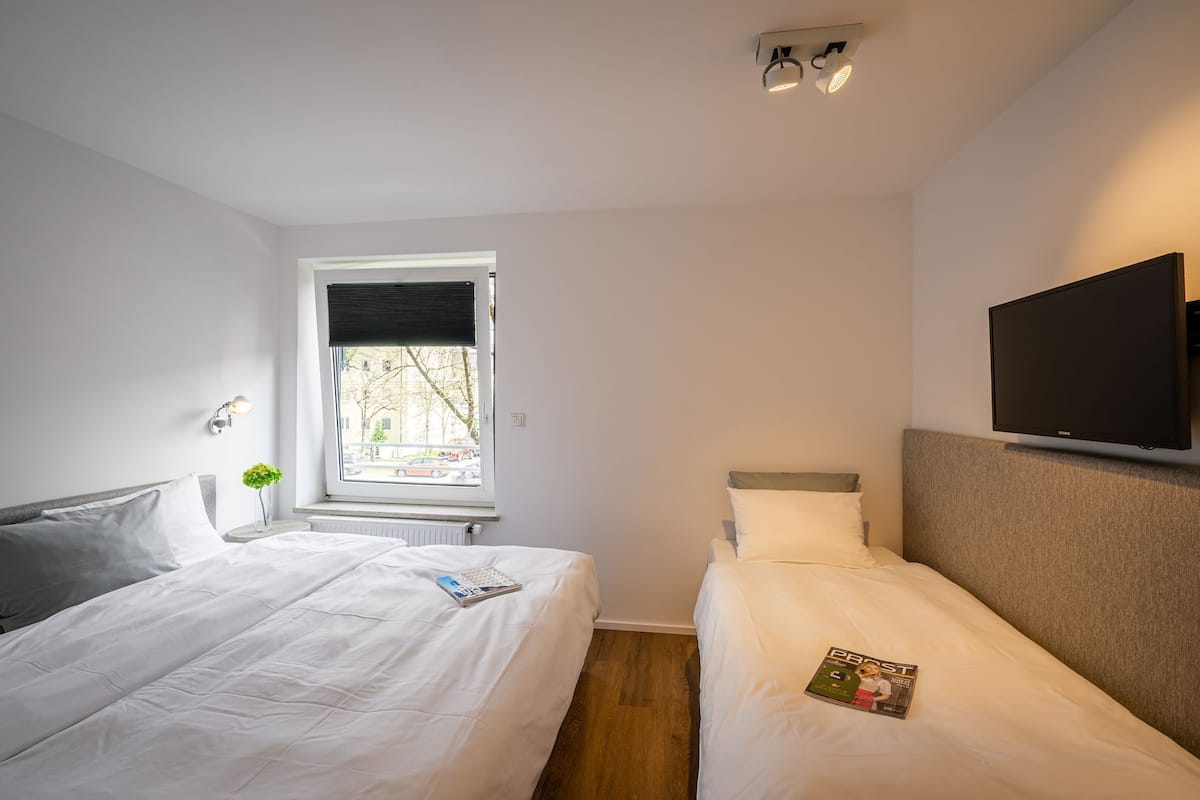 A hotel room with two beds, a window with blinds, a wall-mounted TV, and a radiator.
