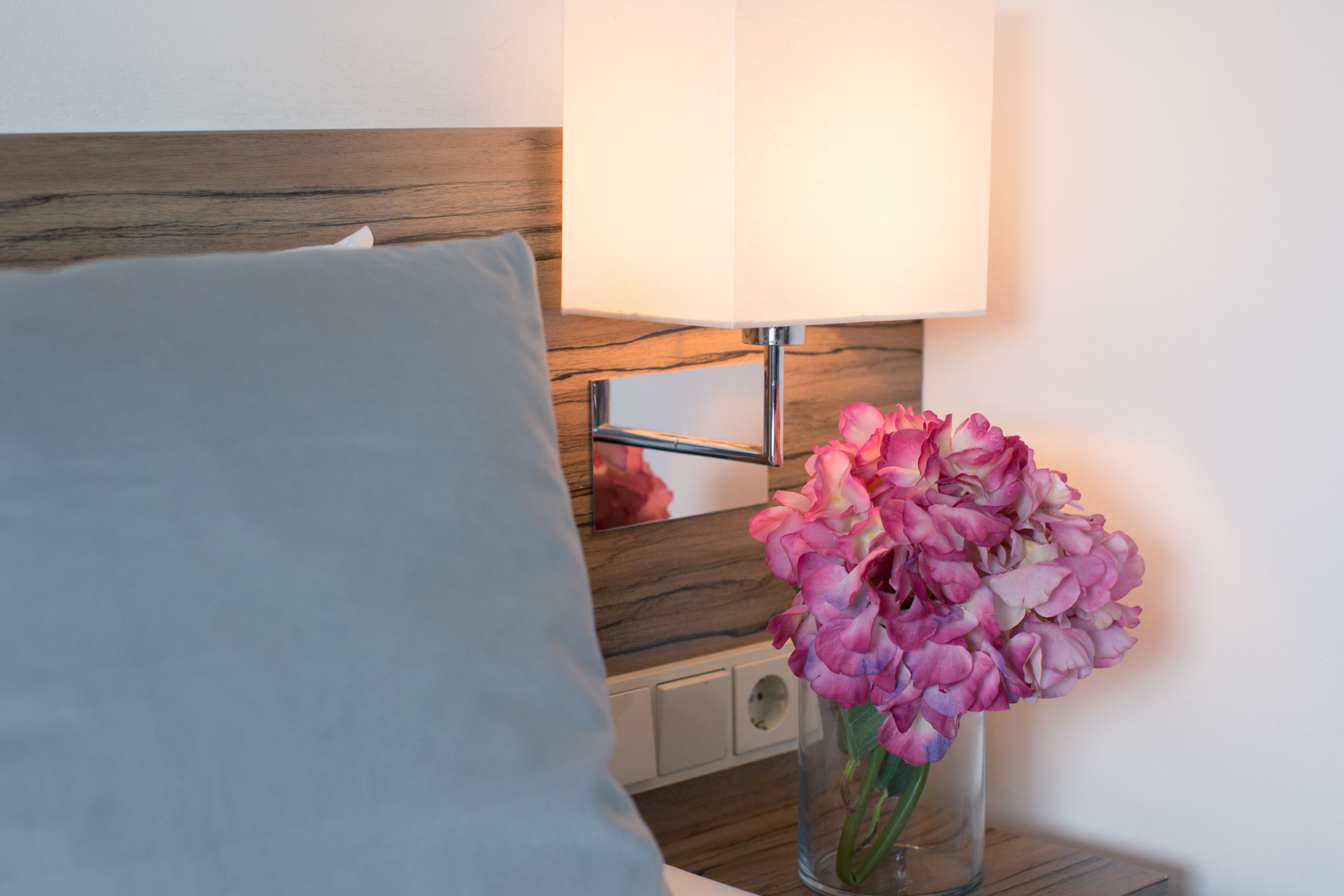 Uma cama com travesseiro cinza, uma mesinha de cabeceira com luminária e um vaso com flores cor-de-rosa.