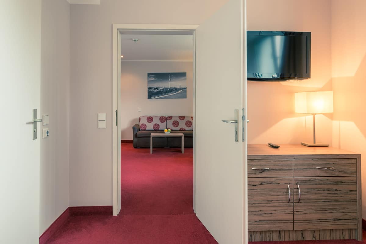 A hotel room with a red carpet, a sofa with a patterned cover, a small table with fruit, a wooden cabinet with drawers, and a mounted TV.