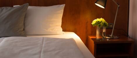 Cama bem arrumada com lençóis e almofadas brancos, mesa de cabeceira com luminária e um vaso de flores, e cabeceira de madeira.