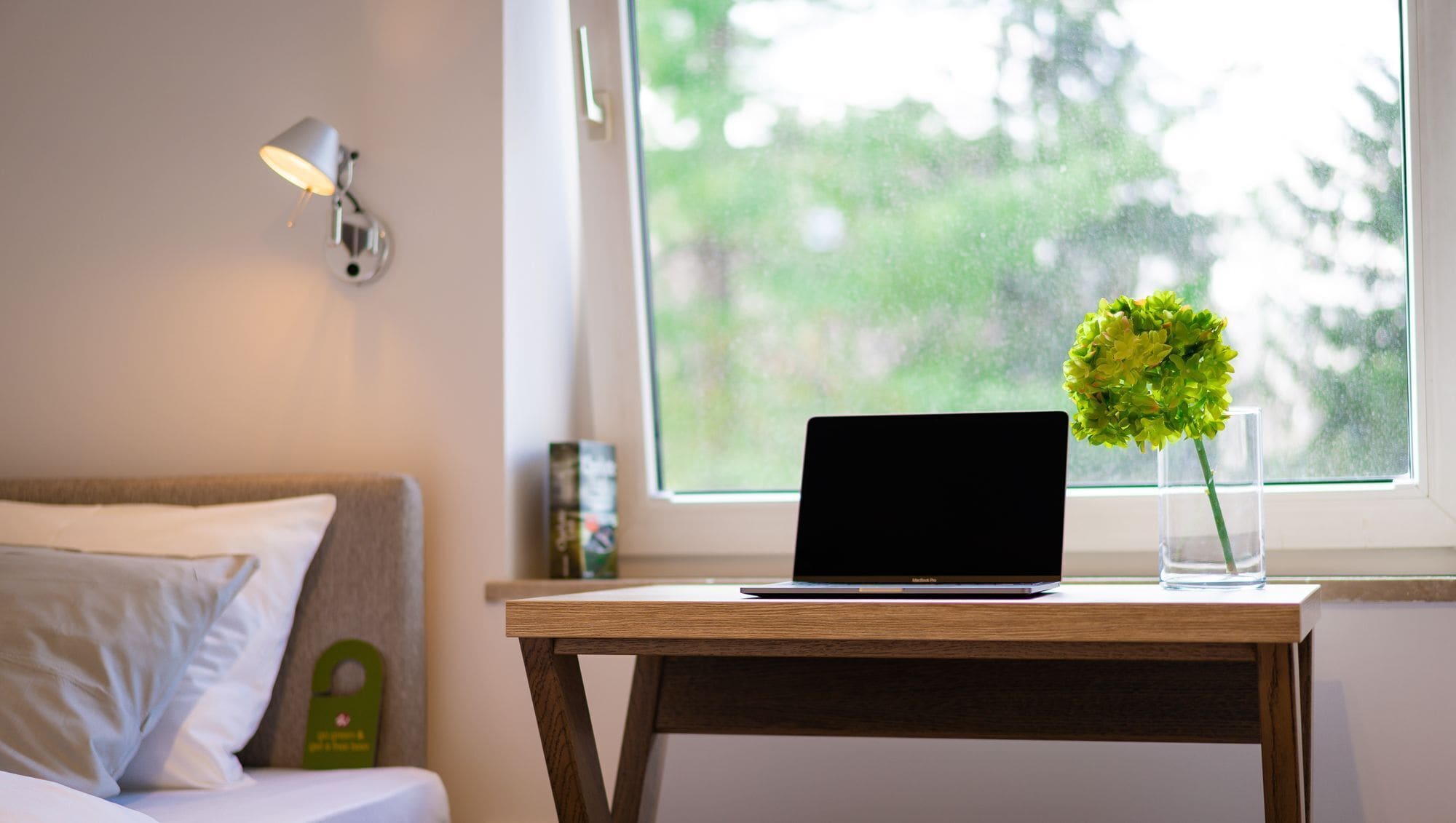 A laptop on a wooden table next to a window with a view of trees.