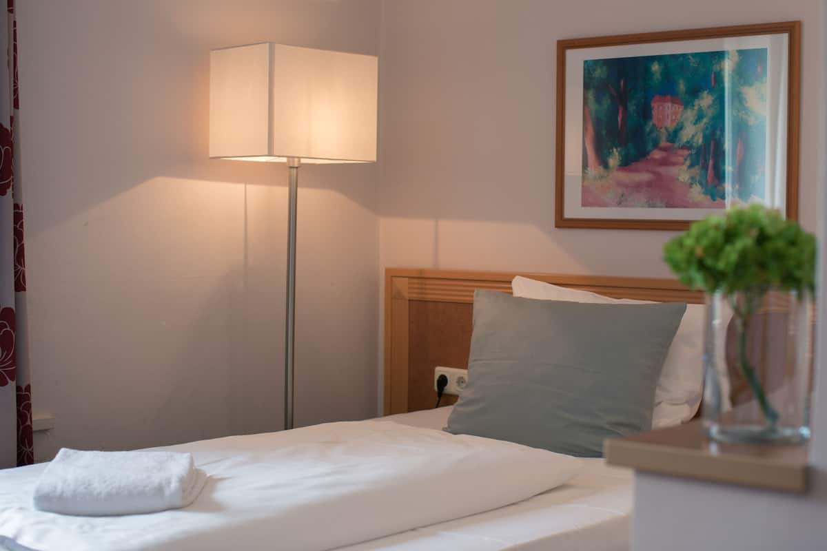 A neatly made bed with a white blanket and a folded towel, a bedside lamp, a framed painting on the wall, and a small potted plant on the nightstand.