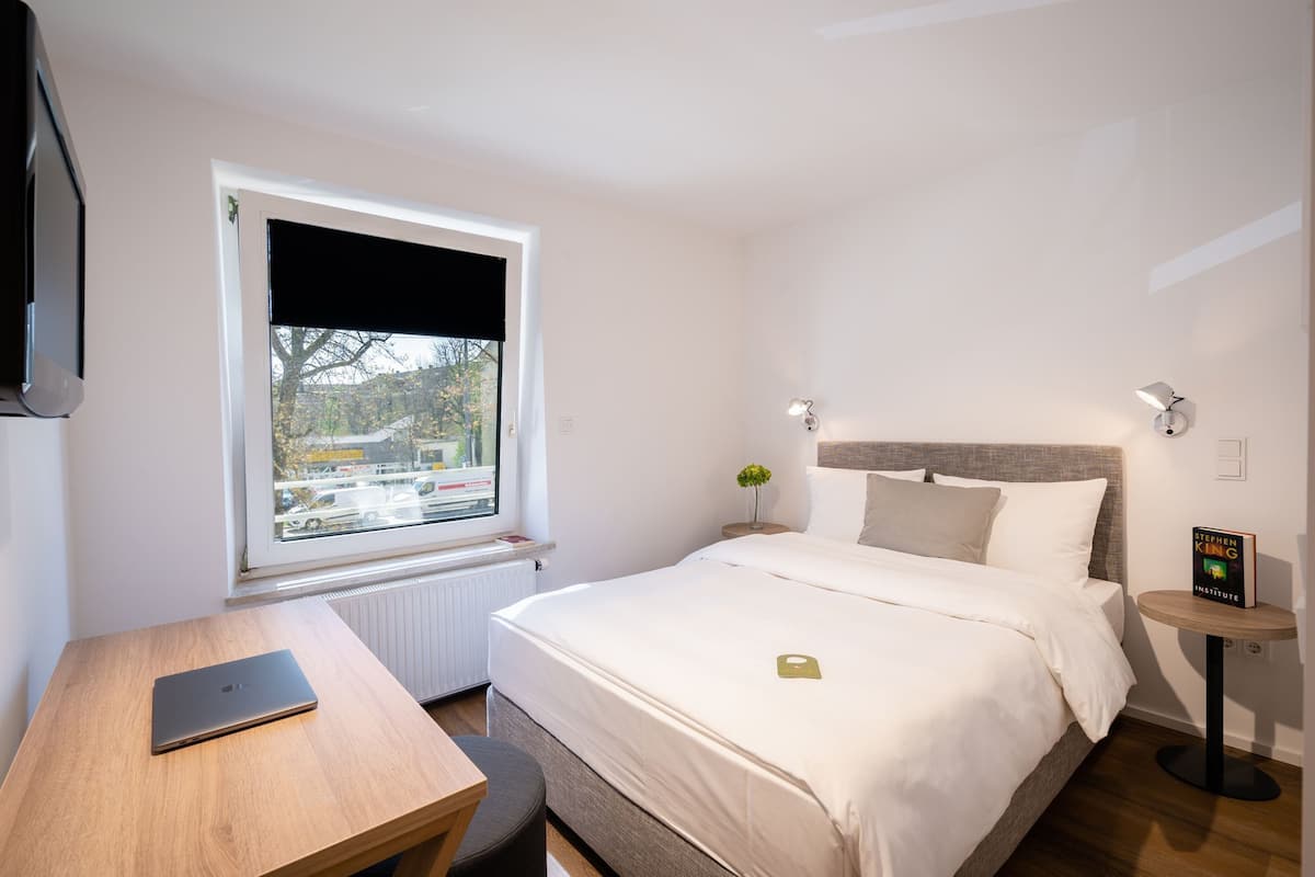 A hotel room with a bed, a desk with a laptop, a window with a view of the street, and a bedside table with a book.