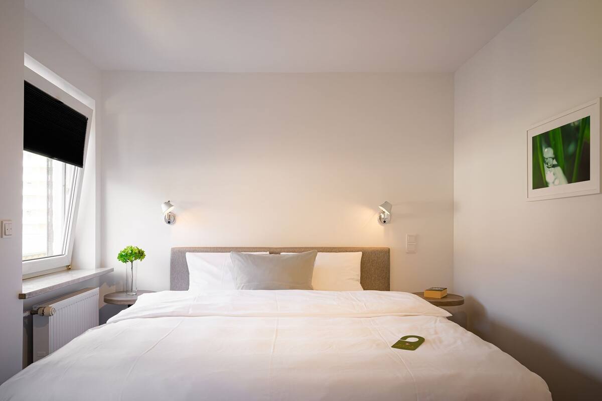 A hotel room with a large bed, two bedside tables, a window with blinds, and a wall-mounted picture.
