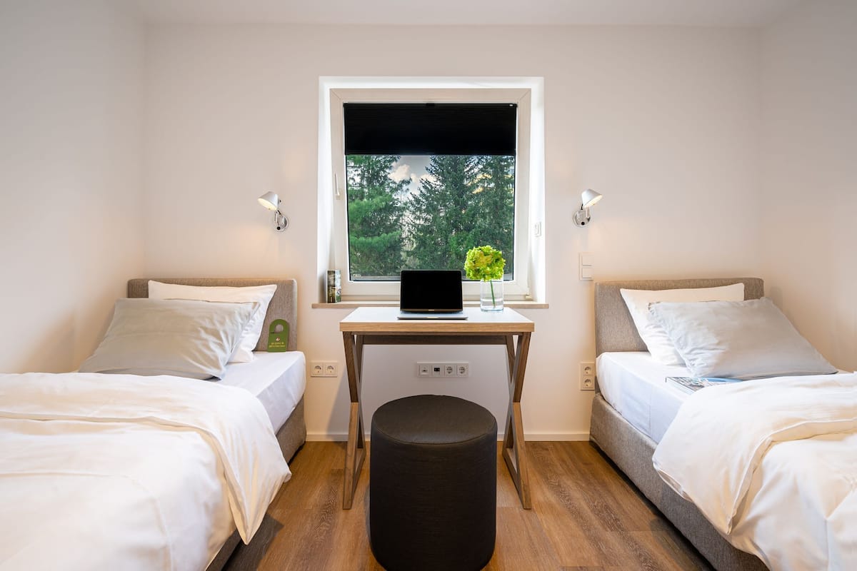 A small, well-lit room with two beds, a desk with a laptop, and a window with a view of trees.