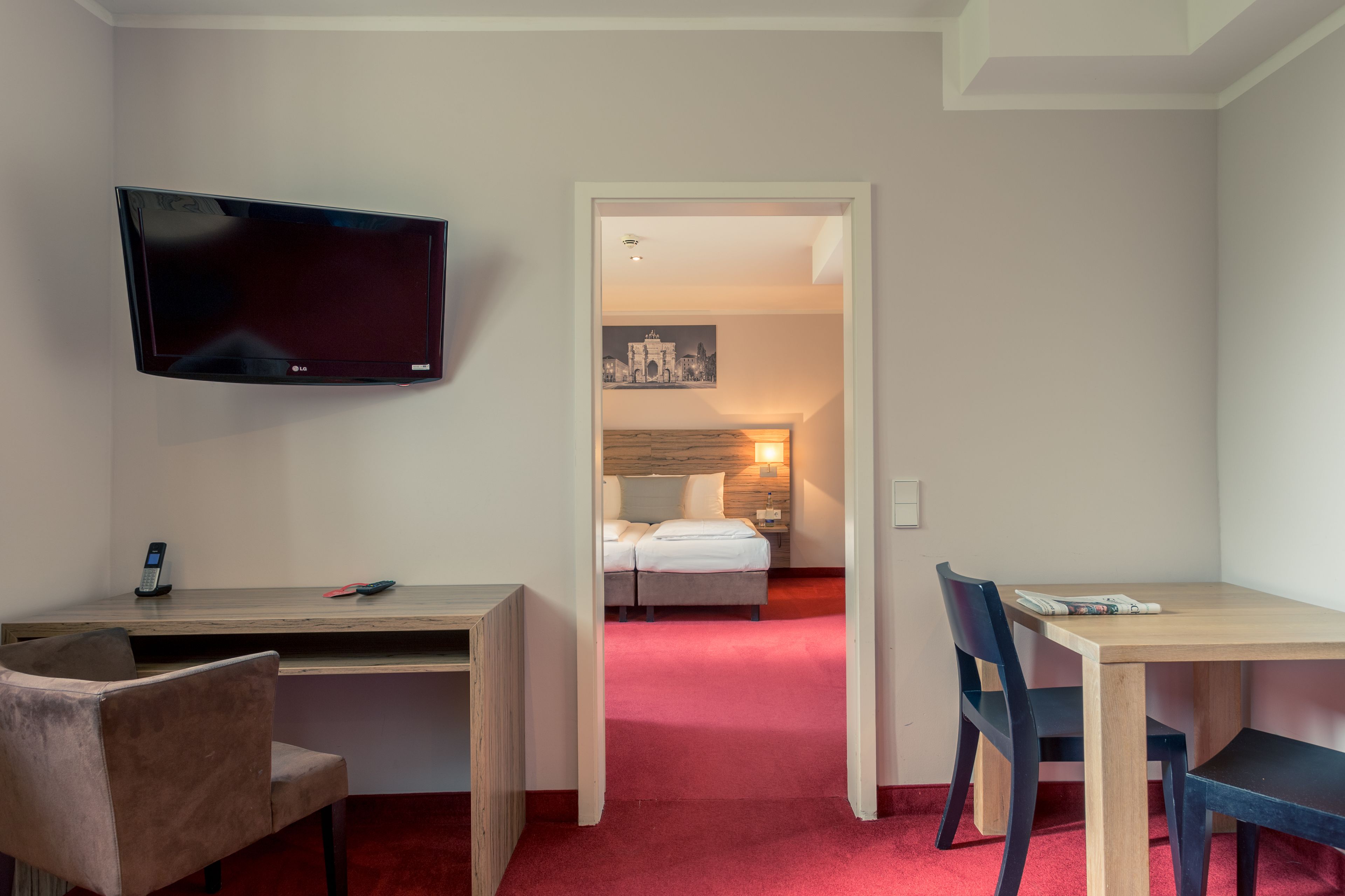 A hotel room with a TV mounted on the wall, a desk with a chair, a bed with a nightstand, and a painting on the wall.