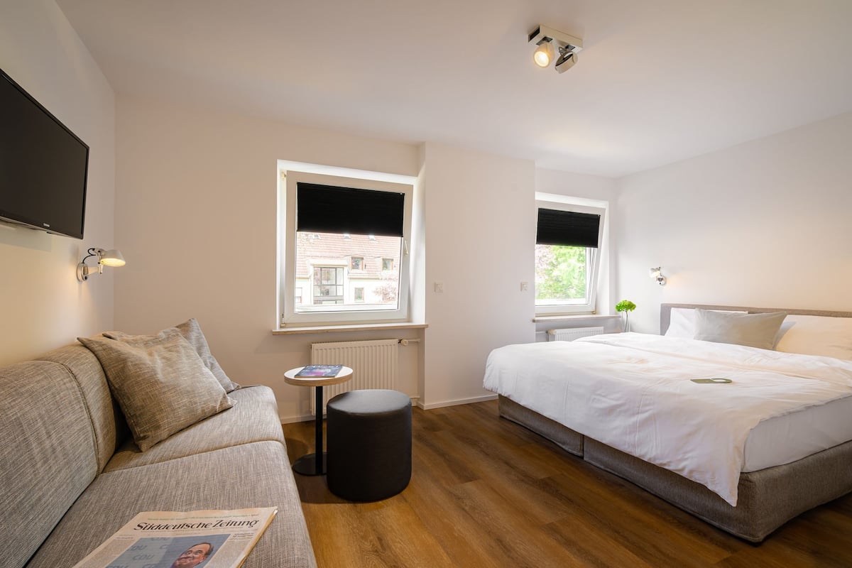 A modern hotel room with a sofa, a small table, a bed, and a window with a view of buildings outside.