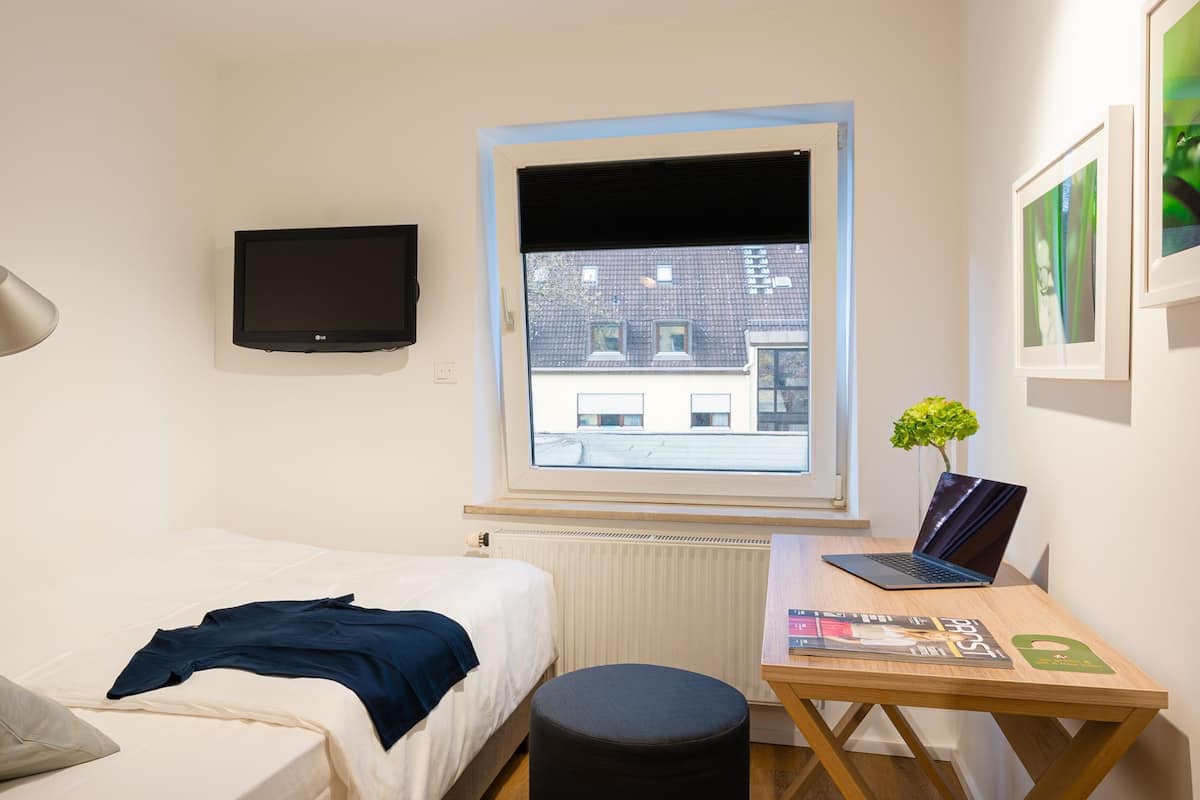A small, well-lit room with a bed, a desk with a laptop, a window with a view of buildings, and a mounted TV.