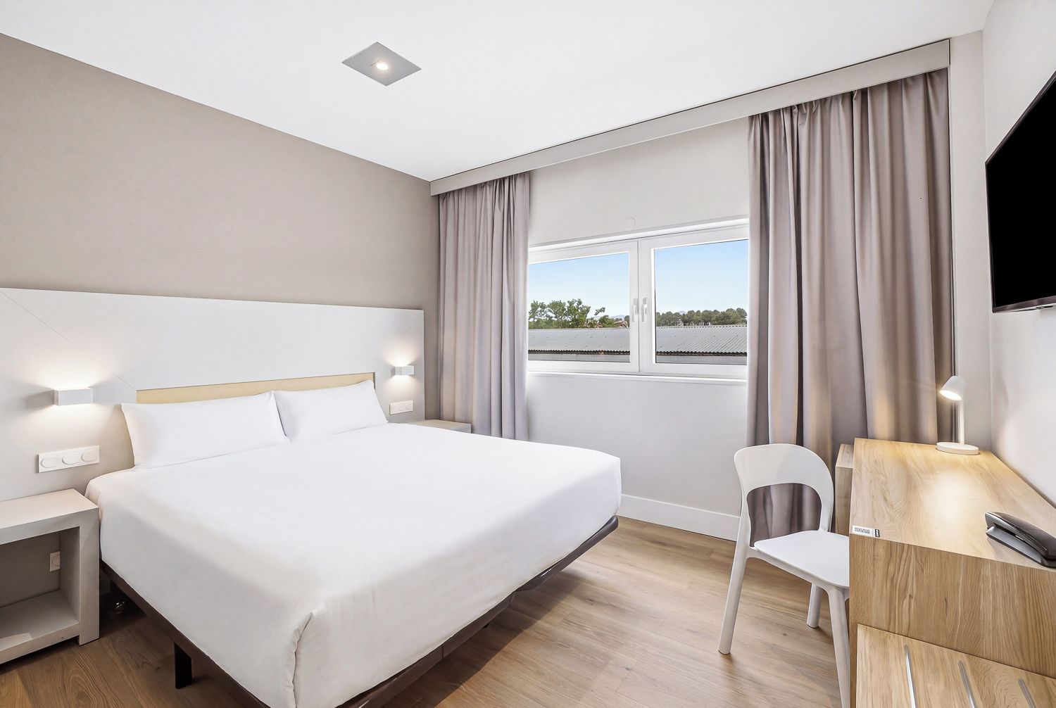 A hotel room with a large bed, a desk with a chair, a TV, and a window with curtains.