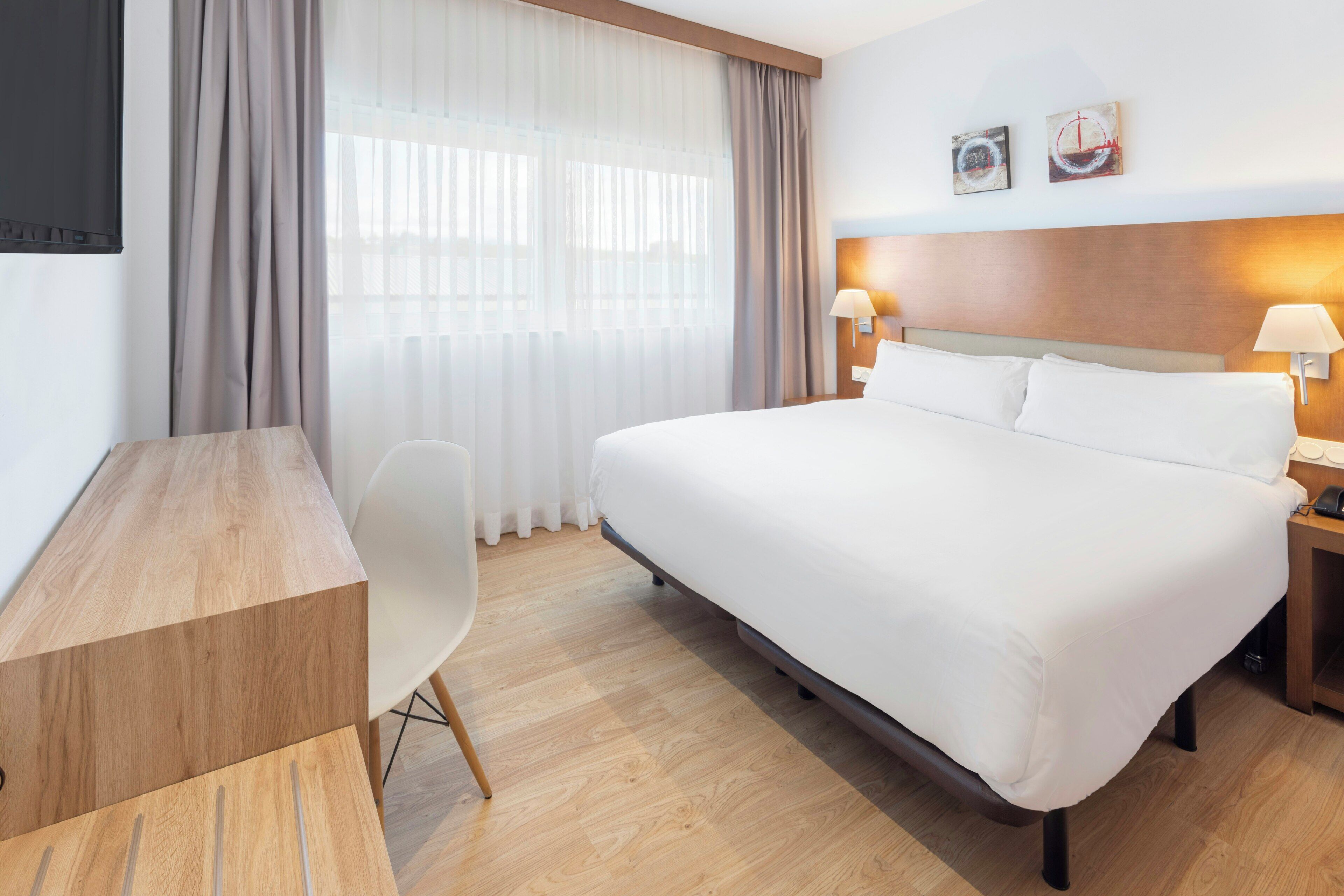 Una habitación de hotel con cama, mesitas de noche, escritorio, silla y ventana con cortinas.