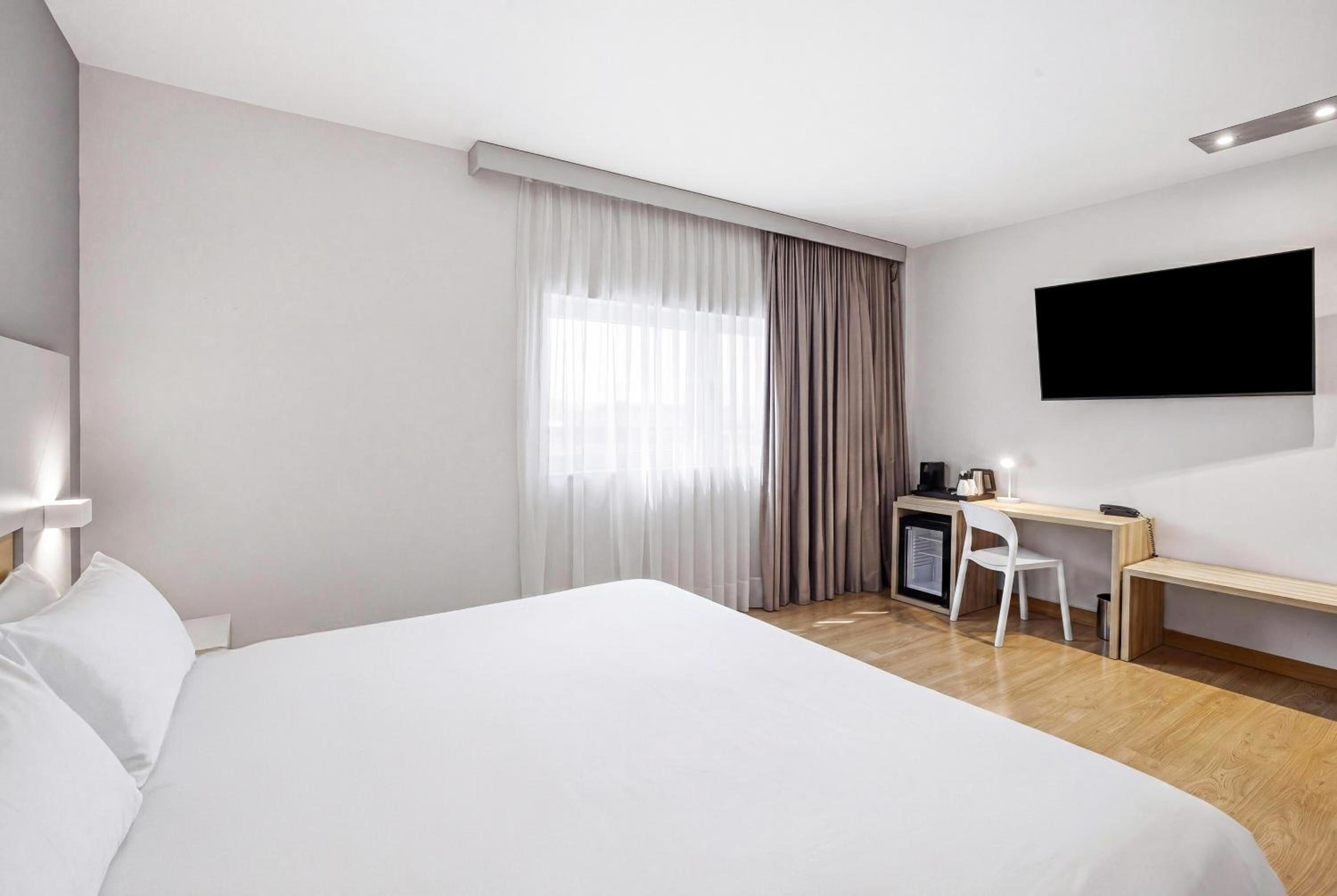 Habitación de hotel con una cama grande, un escritorio, una silla, un televisor y una ventana con cortinas.