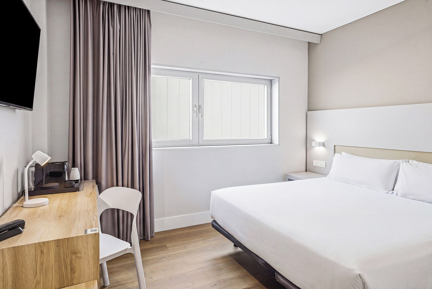A hotel room with a bed, a desk with a chair, a TV, and a window with curtains.