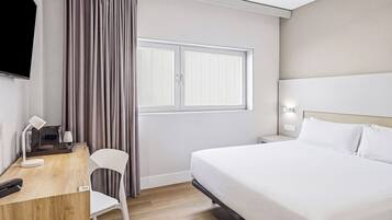 A hotel room with a bed, a desk with a chair, a TV, and a window with curtains.