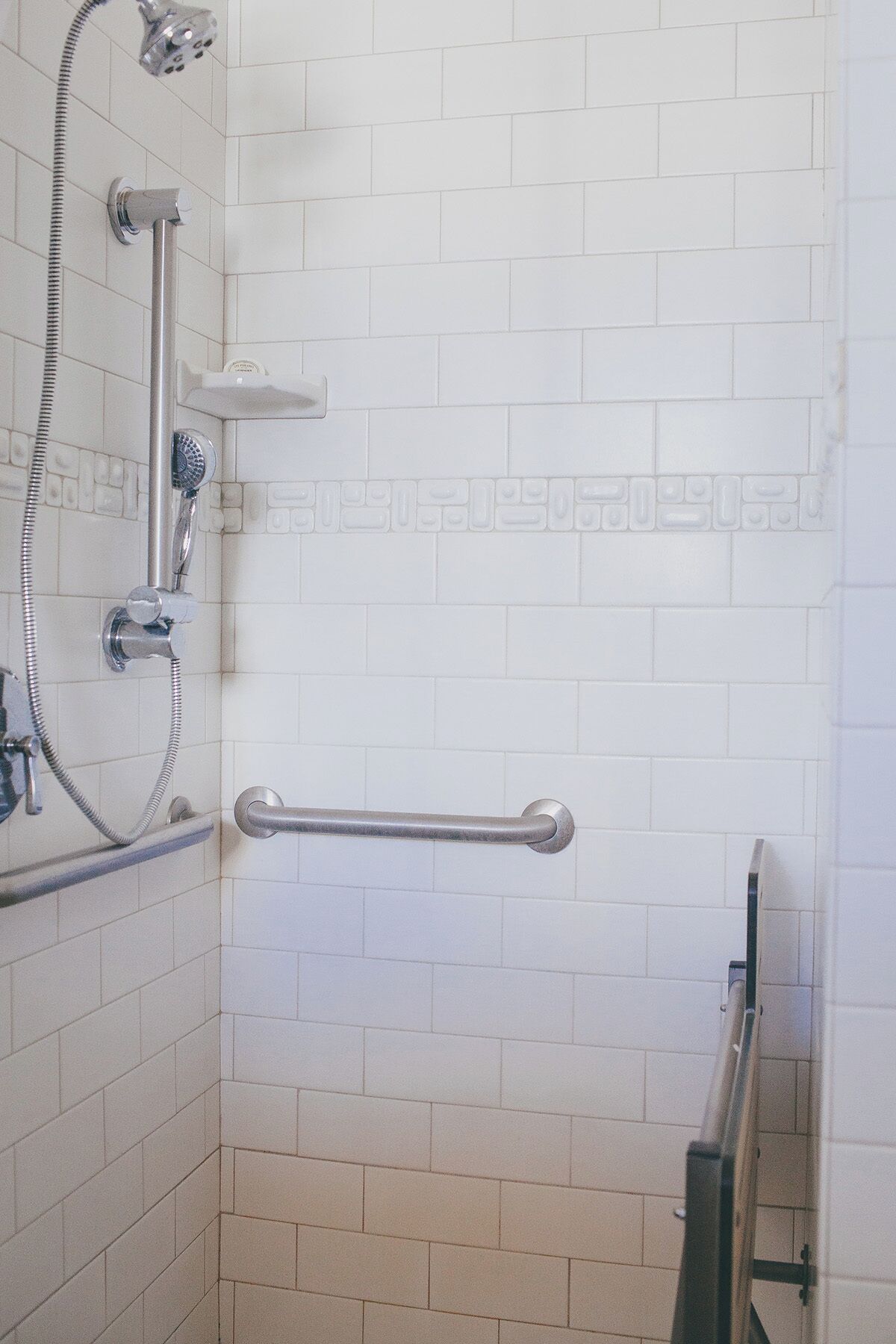 Un baño con paredes de azulejos blancos, una ducha con cabezal de ducha manual y una barra de apoyo.