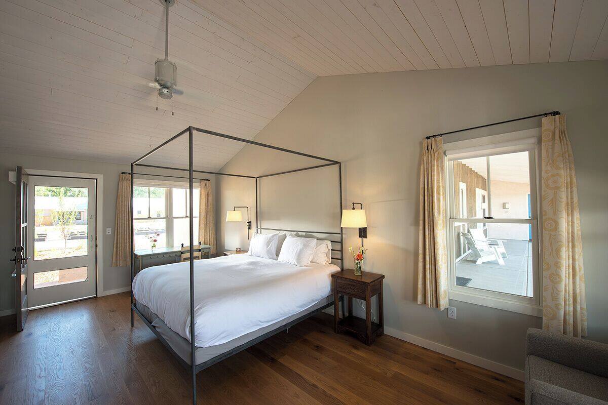 A four-poster bed with a wooden floor, a bedside table with a lamp, and a window with curtains.