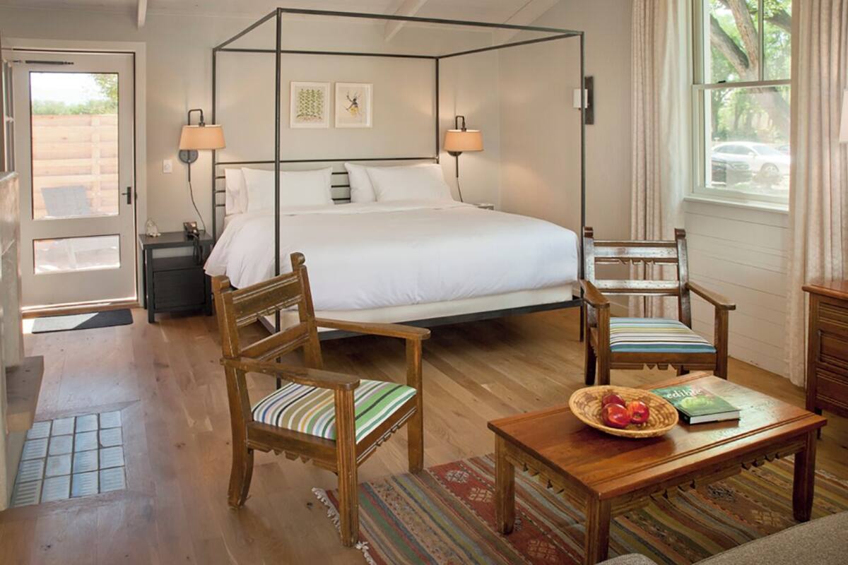 A four-poster bed with white linens, two wooden chairs with striped cushions, a wooden side table with a book and fruit bowl, and a window with curtains.