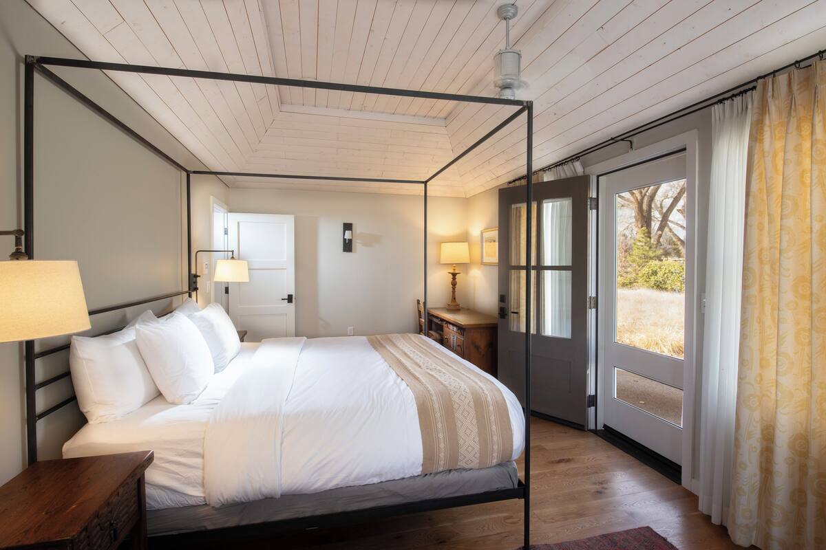 A four-poster bed with white linens, a wooden nightstand, and a bedside table with a lamp. There is a door leading to a balcony with a view of trees and grass.