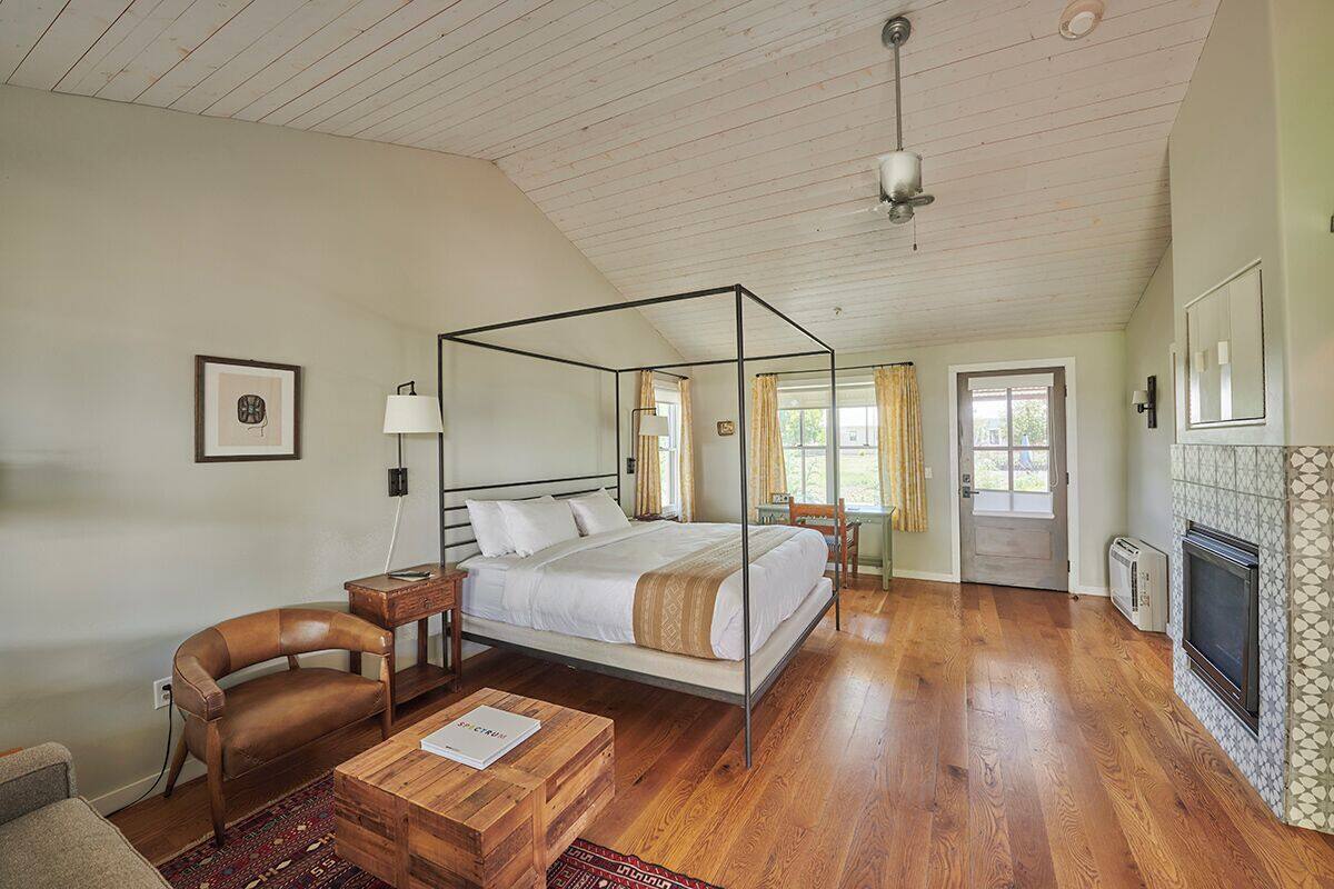 A spacious bedroom with a four-poster bed, a wooden floor, a fireplace, and a view of the outdoors.