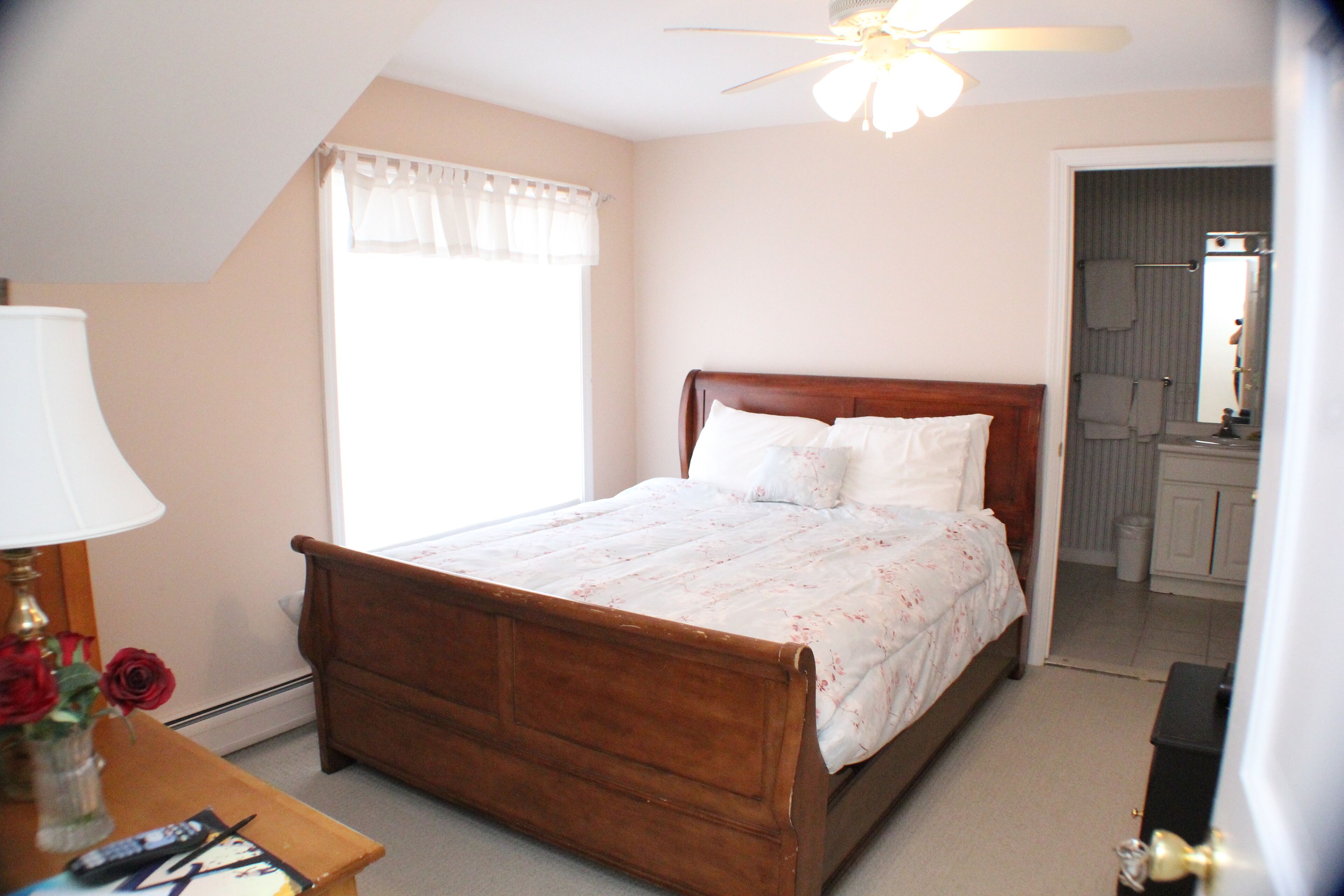 A bedroom with a wooden bed, a nightstand with a lamp and a vase of flowers, a ceiling fan, and a view of the bathroom through an open door.