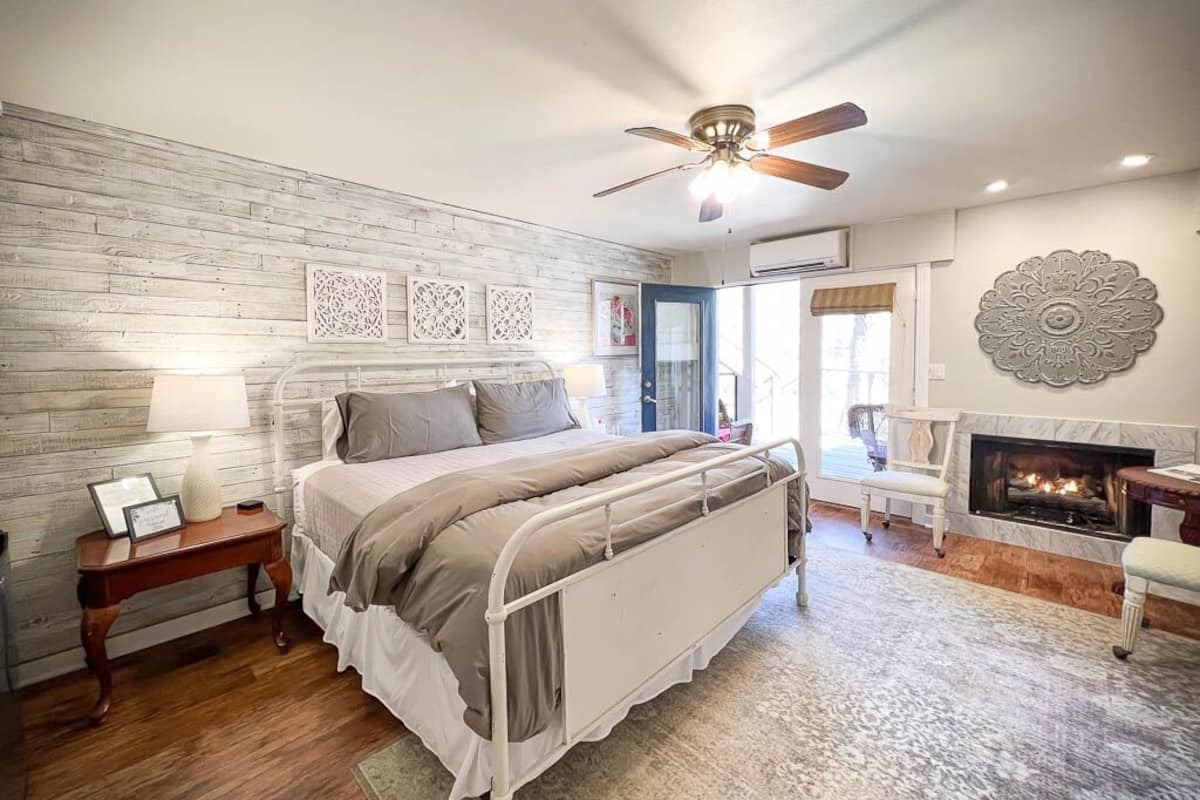 A bedroom with a bed, bedside table, lamp, decorative wall art, a fireplace, and a ceiling fan.
