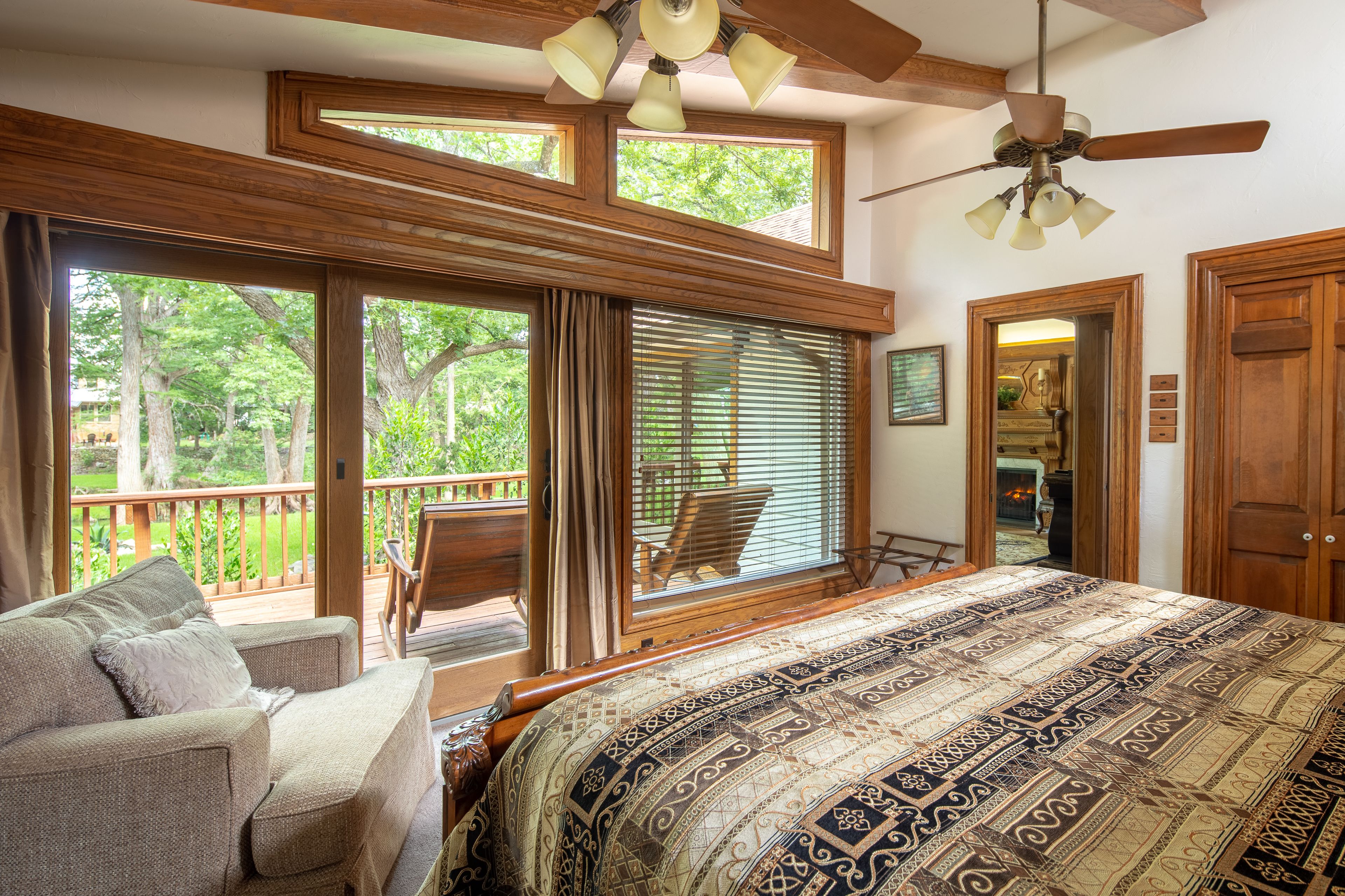 A bedroom with a large bed, a wooden ceiling fan, and a view of a deck and trees outside.