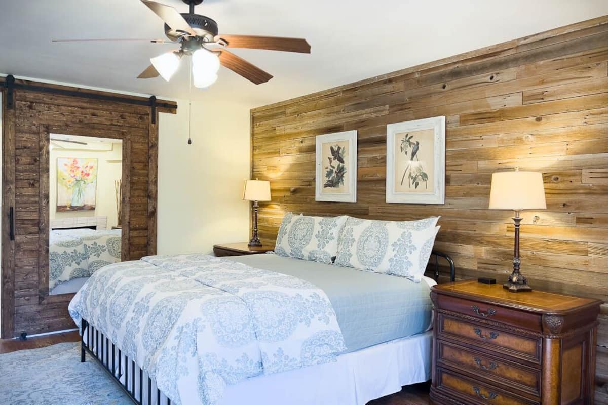 A bedroom with a wooden wall panel, a bed with patterned bedding, two bedside lamps, and a nightstand with drawers.