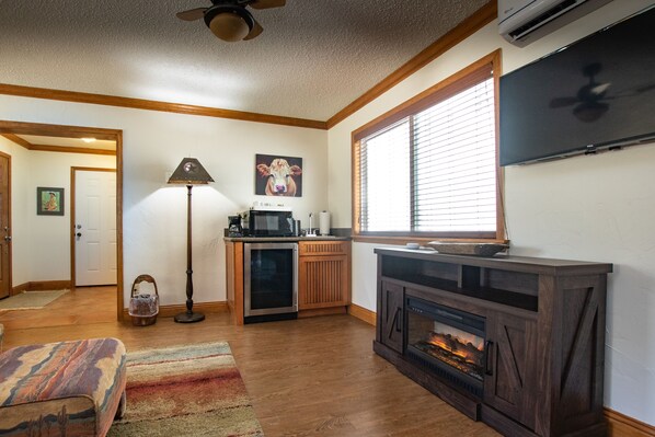 A living room with a fireplace, a television, a kitchen area, and a large window with blinds.