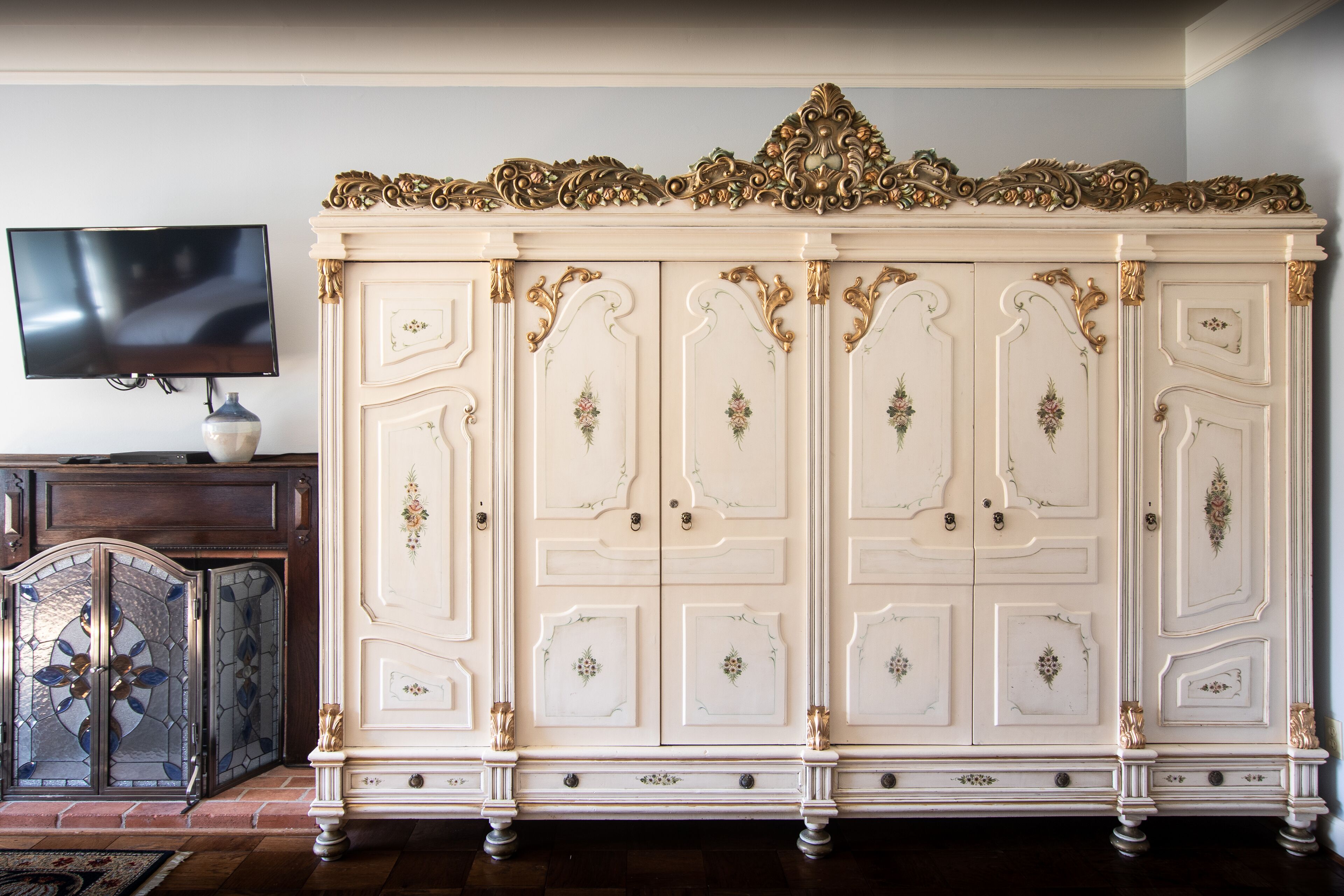 Un'ampia e decorata credenza bianca con dettagli dorati e motivi floreali, posizionata accanto a un televisore a schermo piatto su un mobile in legno.