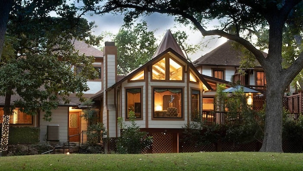 A two-story house with a prominent attic window and a large tree in the front yard. - Creekhaven Inn & Spa (Wimberley)