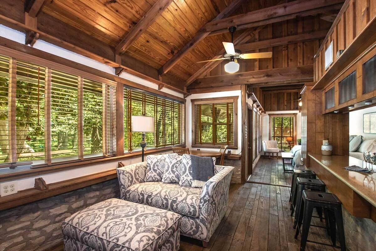 A wooden interior with a sofa and ottoman, large windows, and a ceiling fan.