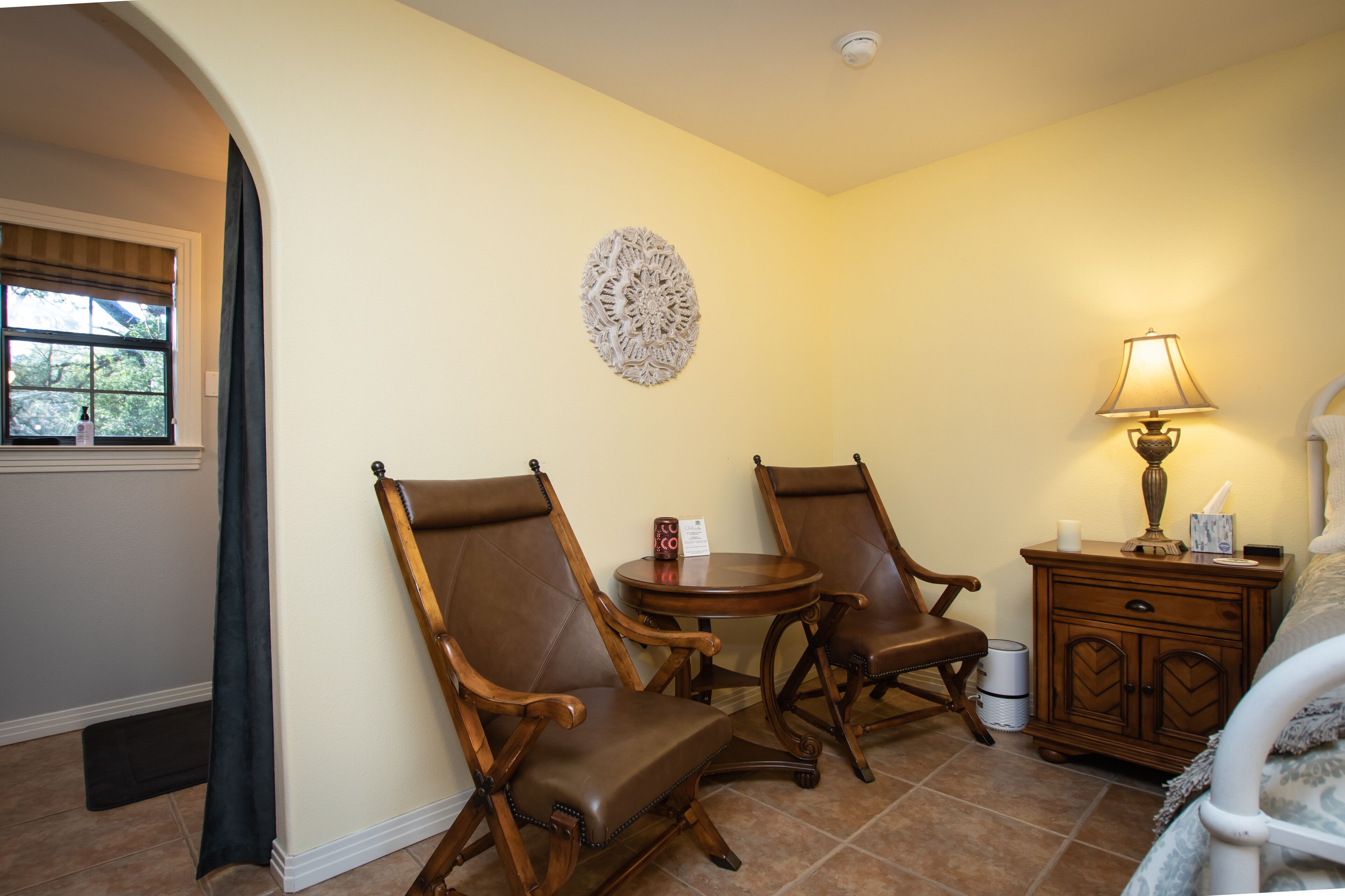 A room with two leather chairs, a small round table, a lamp, a nightstand, and a bed.