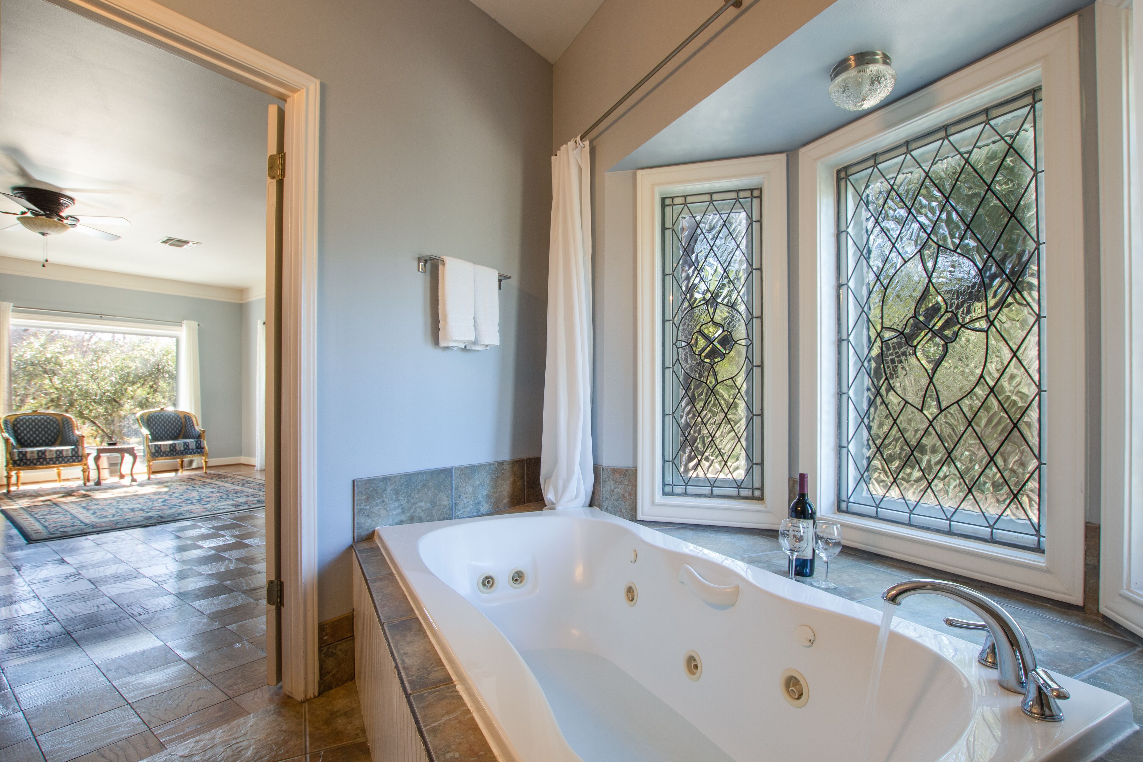 Une salle de bain spacieuse avec une grande baignoire, une fenêtre ornée de motifs décoratifs en verre et une vue sur un espace extérieur aménagé pour se détendre.
