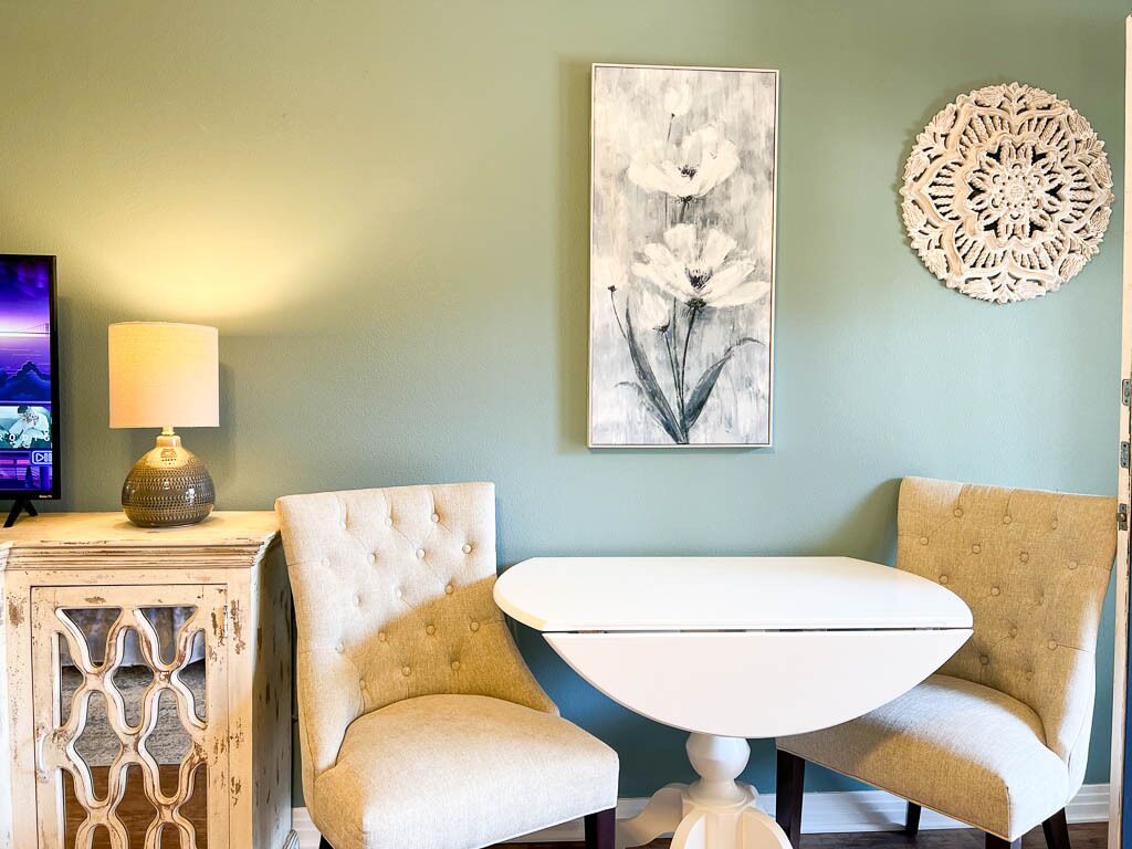Een kamer met een witte tafel en twee beige fauteuils, een decoratief wandobject en een lamp op een bijzettafeltje.
