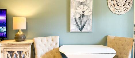 Een kamer met een witte tafel en twee beige fauteuils, een decoratief wandobject en een lamp op een bijzettafeltje.