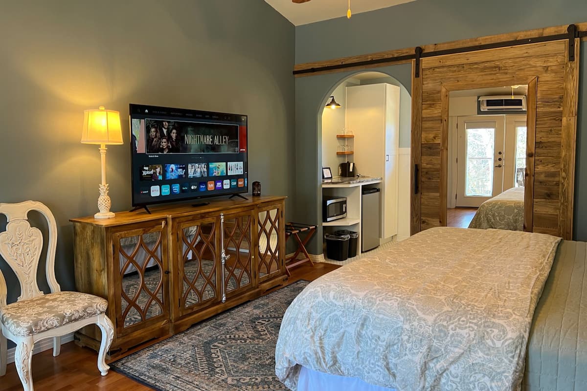 A bedroom with a large bed, a TV on a wooden console, a chair, and a small kitchenette.
