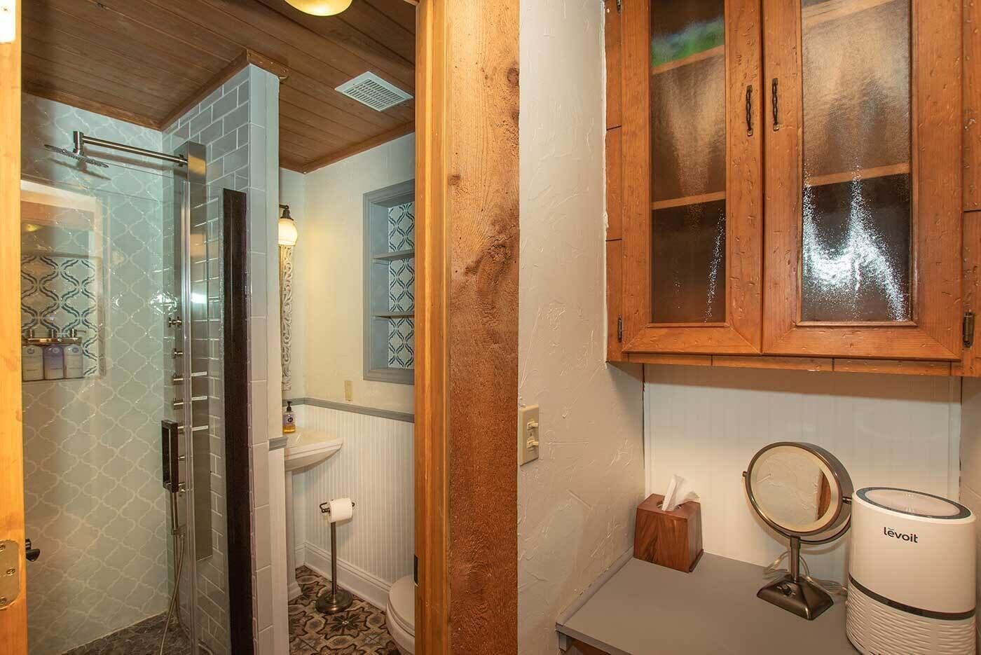 A bathroom with a glass shower enclosure, a wooden cabinet with glass doors, a toilet, and a vanity with a mirror and a Levoit air purifier.