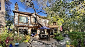 A rustic stone patio with outdoor seating, a large window, and a balcony with an American flag. - Creekhaven Inn & Spa (Wimberley)