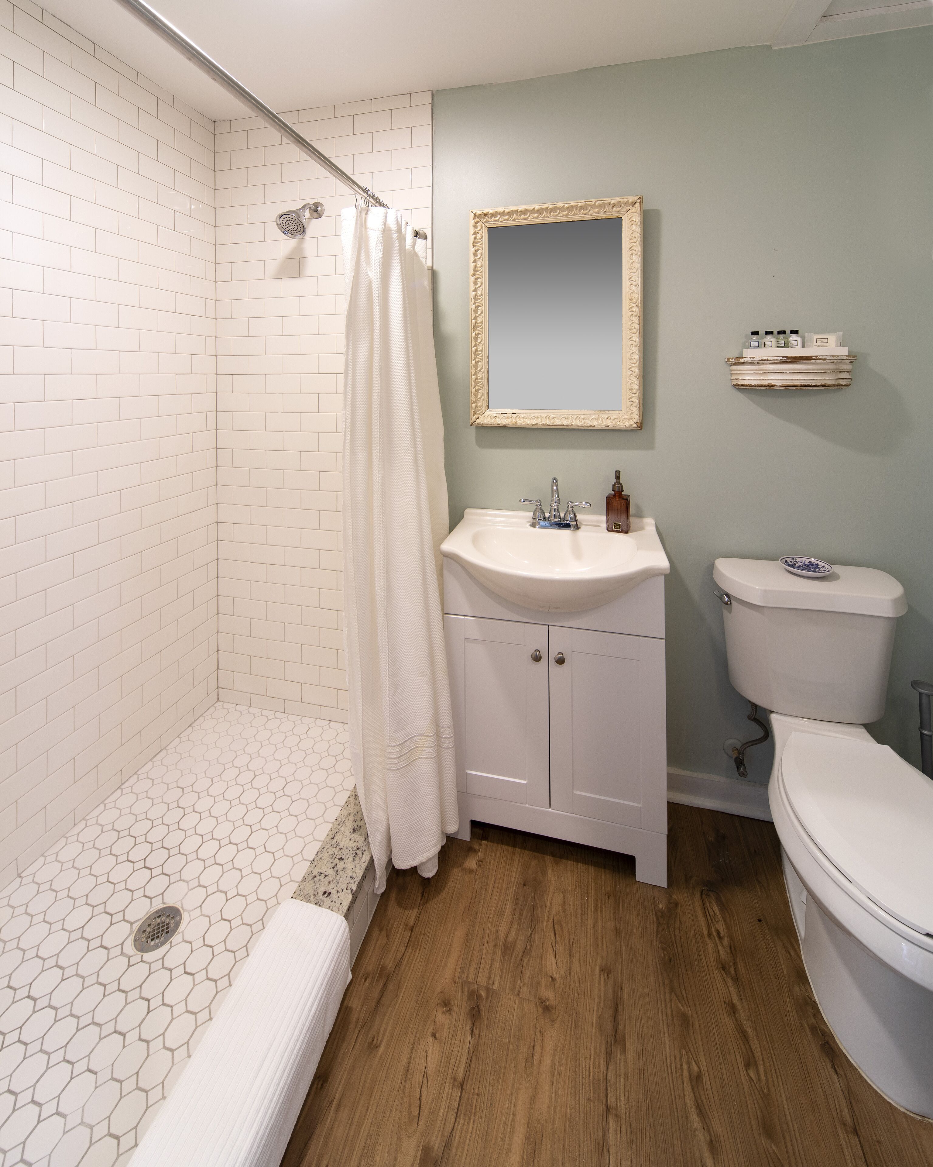 Une salle de bain avec des murs recouverts de carreaux blancs, une douche avec un rideau blanc, un lavabo avec un miroir au-dessus et des toilettes.