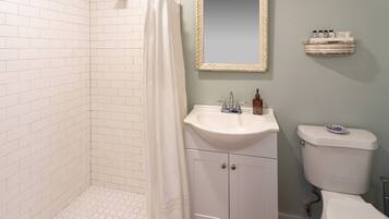 Une salle de bain avec des murs recouverts de carreaux blancs, une douche avec un rideau blanc, un lavabo avec un miroir au-dessus et des toilettes.