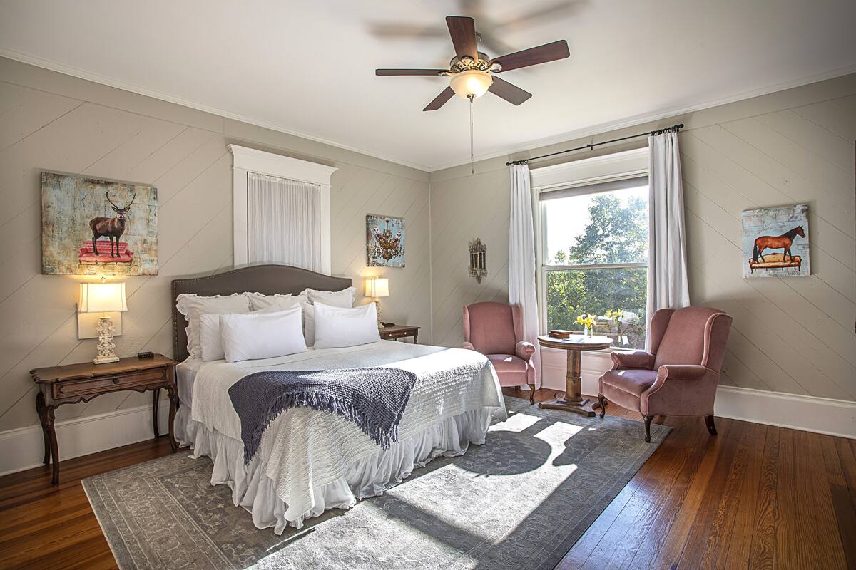 Un dormitorio con una cama grande, un ventilador de techo, una ventana con cortinas, una silla y obras de arte en las paredes.