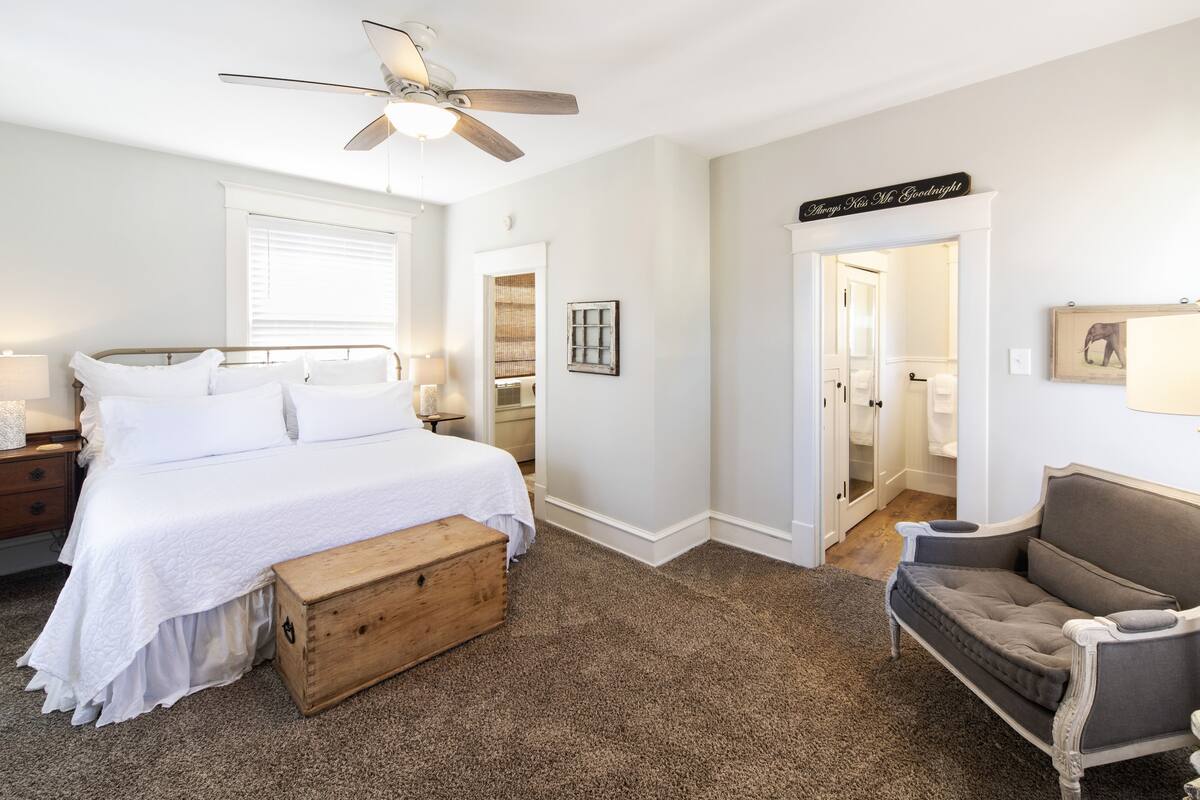 Un dormitorio con cama, un cofre de madera, un sillón gris, un ventilador de techo y un letrero en la pared.