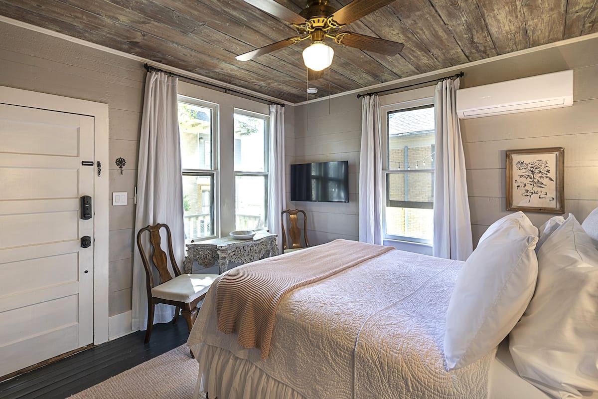 Un dormitorio con una cama grande, un ventilador de techo, un televisor montado en la pared y una ventana con cortinas.