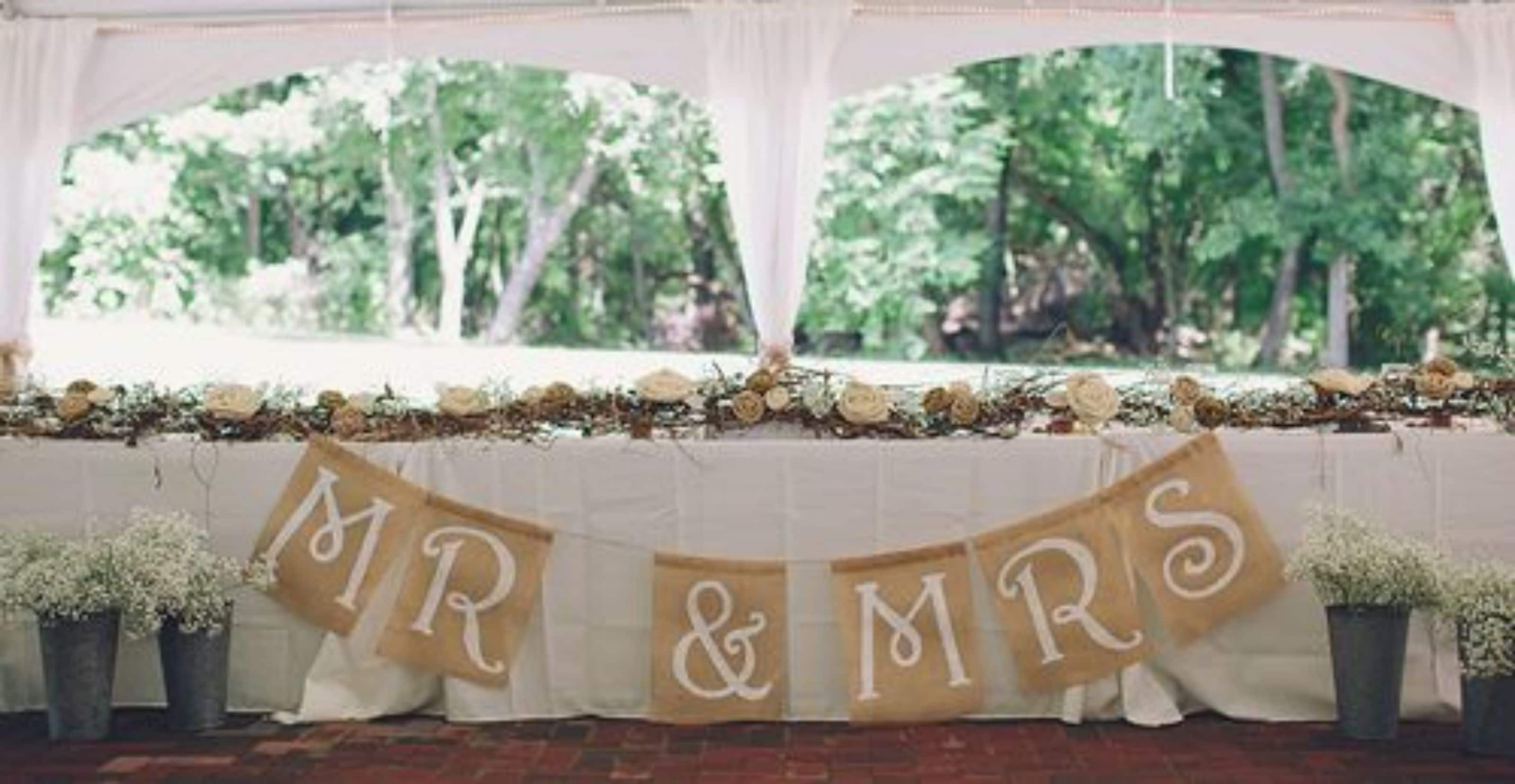 A wedding arch with "MR & MRS" banner, potted plants, and a view of trees through the arch.