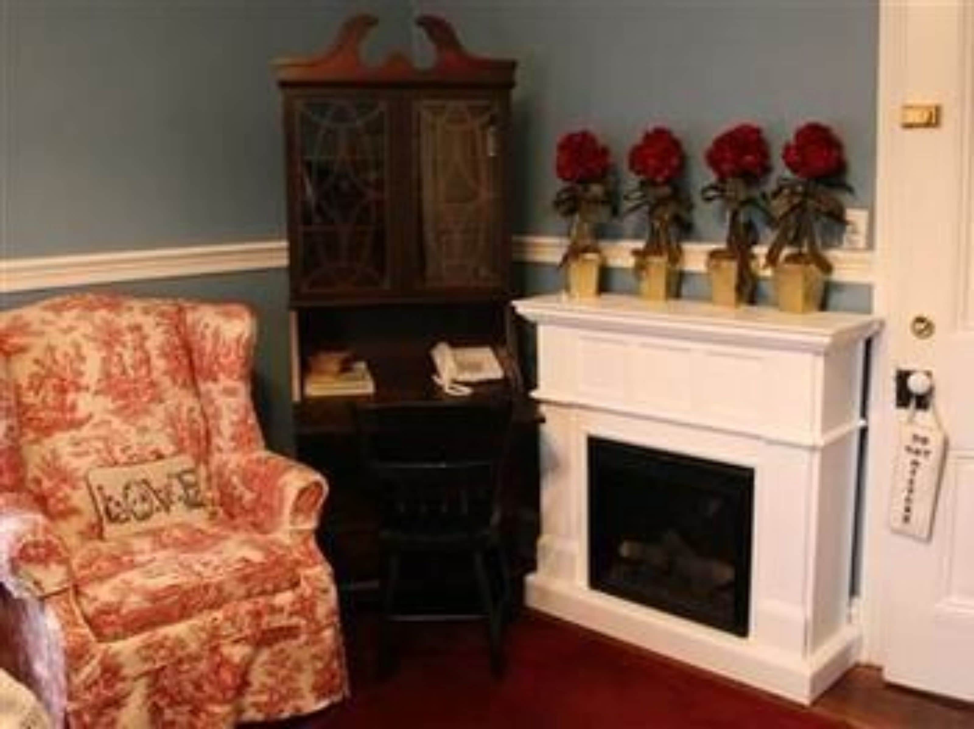 Una habitación con un sillón estampado, un aparador de madera, una chimenea y flores rojas en la repisa.