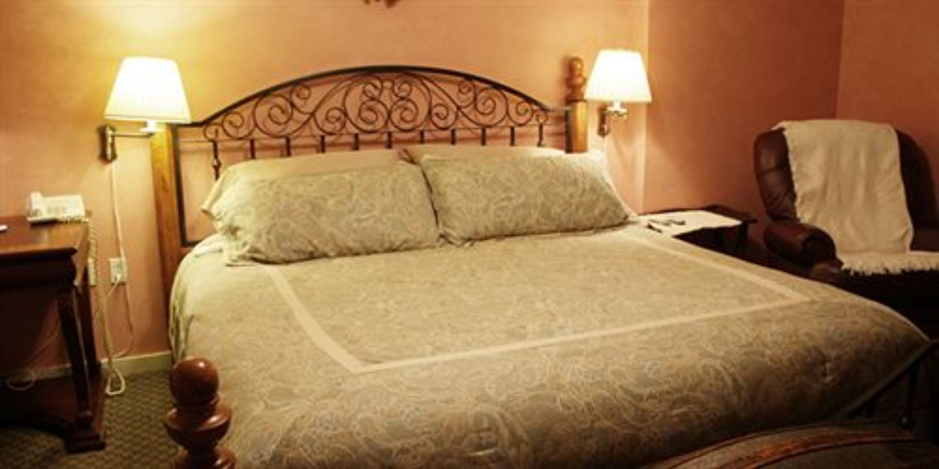 A hotel room with a large bed, two bedside lamps, a wooden nightstand, and a chair with a white cover.