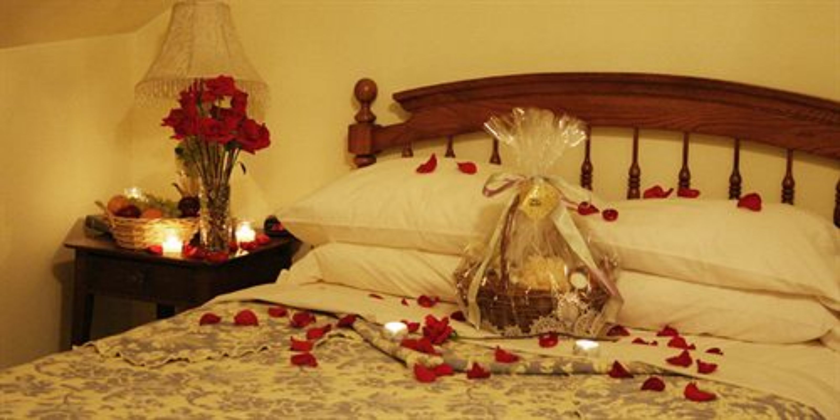 Una cama con pétalos de rosa esparcidos, una mesita de noche con una lámpara y un jarrón de flores rojas, y un objeto decorativo sobre la cama.
