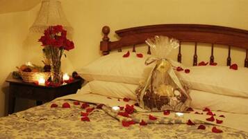 Una cama con pétalos de rosa esparcidos, una mesita de noche con una lámpara y un jarrón de flores rojas, y un objeto decorativo sobre la cama.