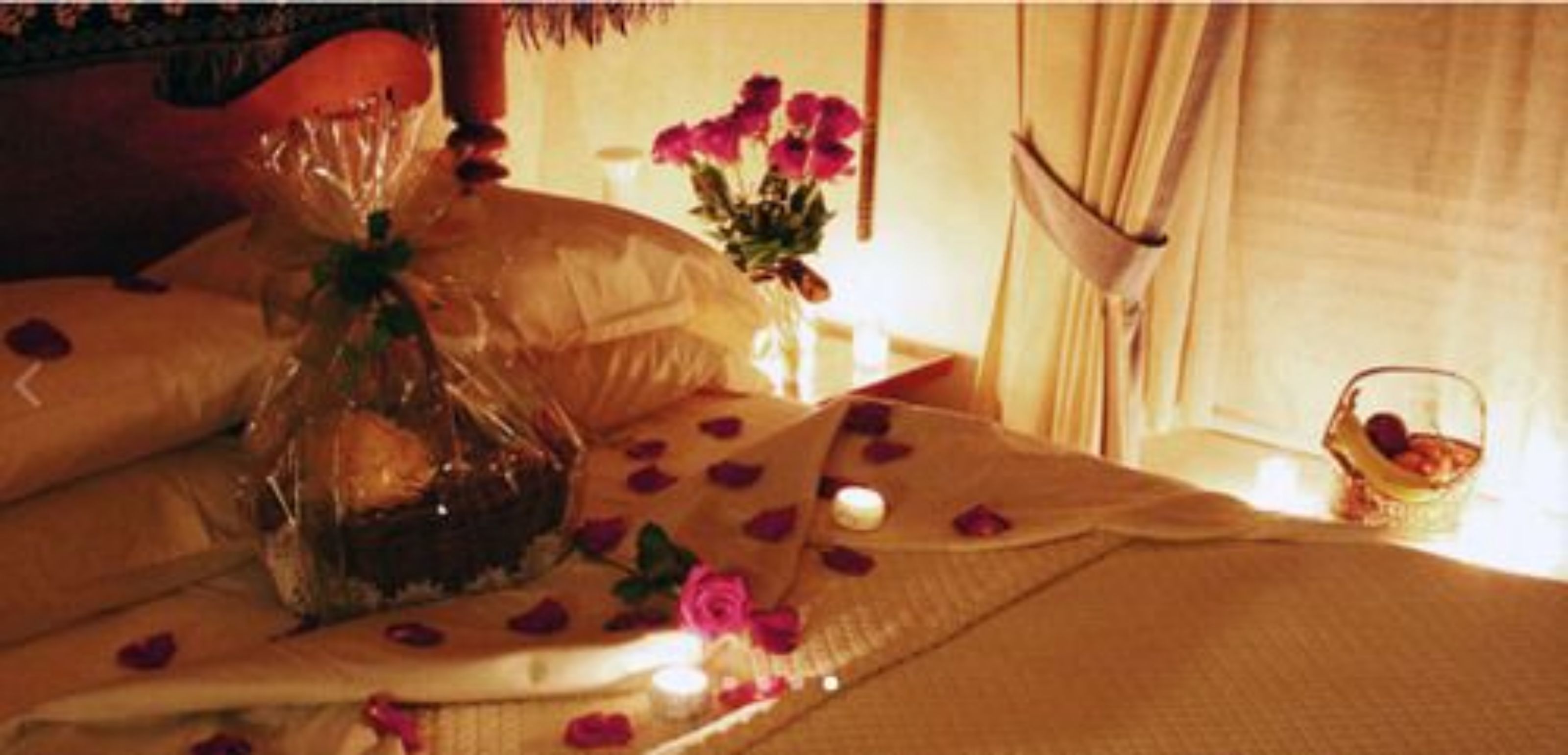A bed with rose petals, a candle, and a basket of fruit.